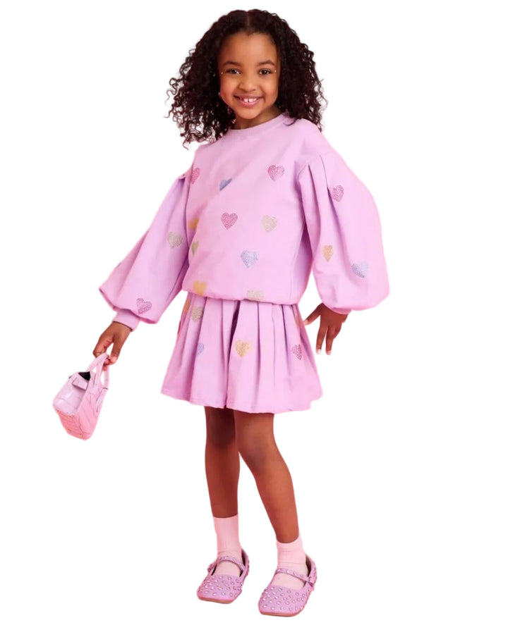 A young girl with curly dark hair stands against a solid pink studio background wearing a light purple/lavender two-piece outfit with colorful heart prints, holding a matching small handbag.