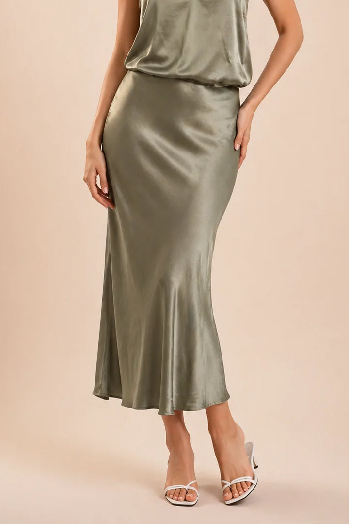 A woman modeling a satin slip dress in a muted sage green color, paired with white heeled sandals, against a solid beige studio background.