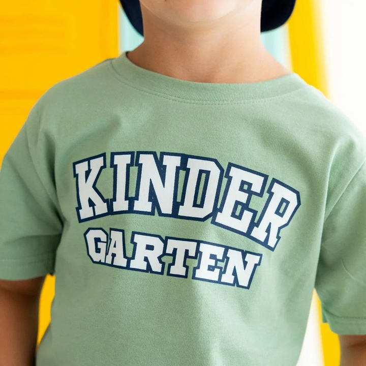 Child wearing Sweet Wink's sage tee with white and navy Kindergarten varsity lettering