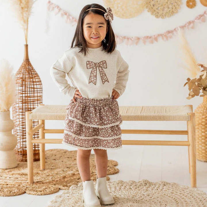 A young girl stands in a styled indoor room wearing a cream long-sleeve sweater with a brown bow graphic and a brown floral tiered skirt with white lace trim, paired with white ankle boots.