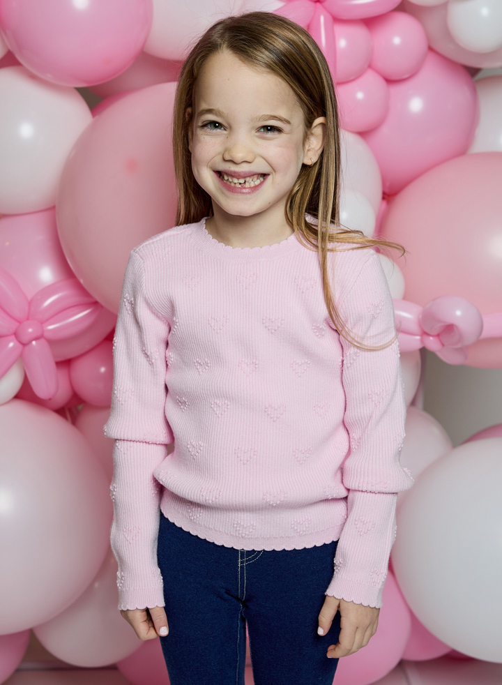 A young girl smiling in front of a pink and white balloon backdrop, wearing a light pink textured knit sweater with dark blue jeans.
