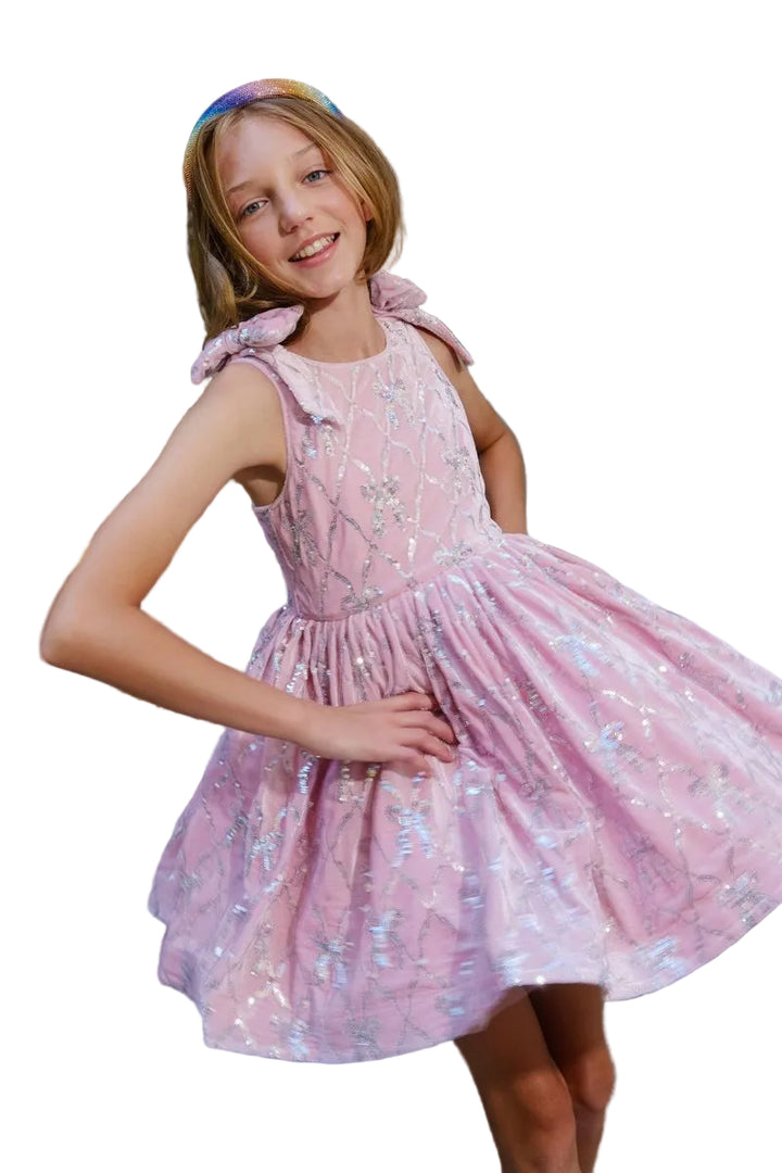 A young girl wearing a pink sleeveless dress with silver sequin patterns and matching pink bow headband, posing on stone steps in front of a building.
