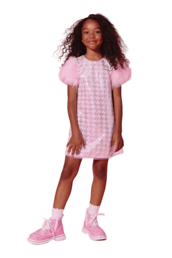 A young girl with curly black hair stands against a solid pink studio background wearing a pink and white patterned dress with fluffy pink sleeves, white socks, and pink sneakers.