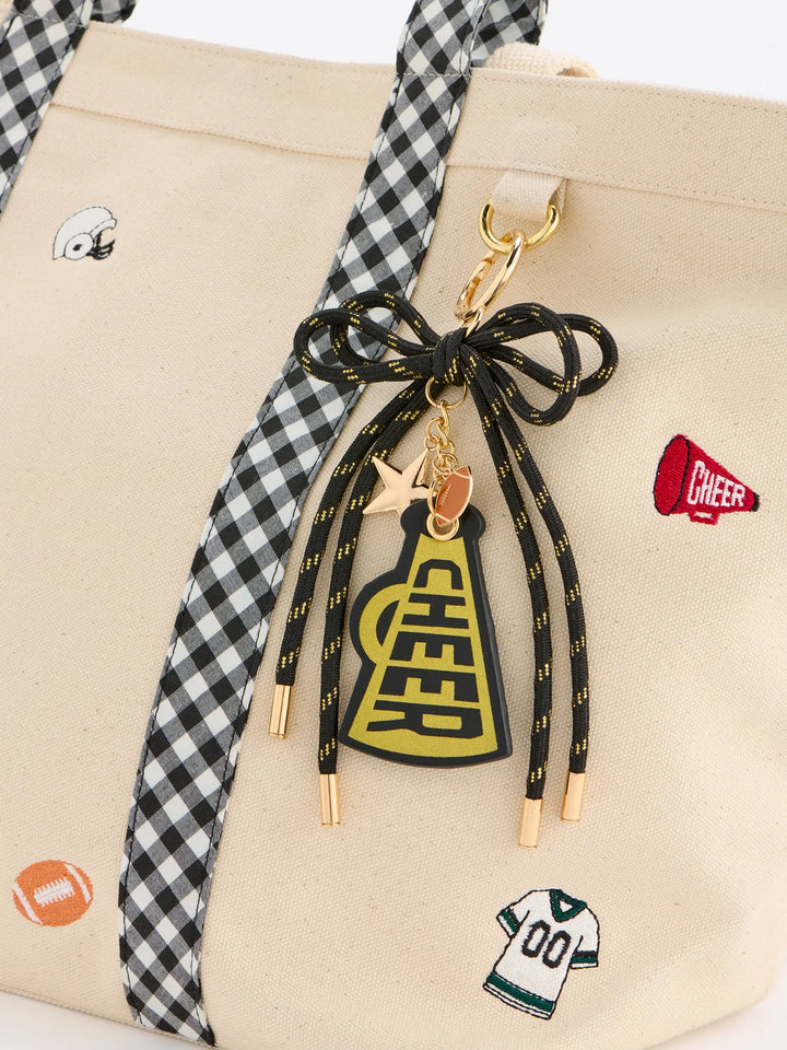 A close-up of a beige tote bag with black-and-white gingham straps, decorated with small sports-themed patches and a large black-and-yellow cheerleader pom-pom keychain charm with gold-tone hardware.
