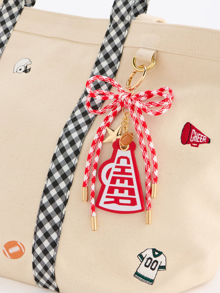 A close-up of a beige tote bag with black-and-white gingham handles, decorated with small sports-themed patches and a red-and-white cheerleader rope charm with gold-tone hardware.