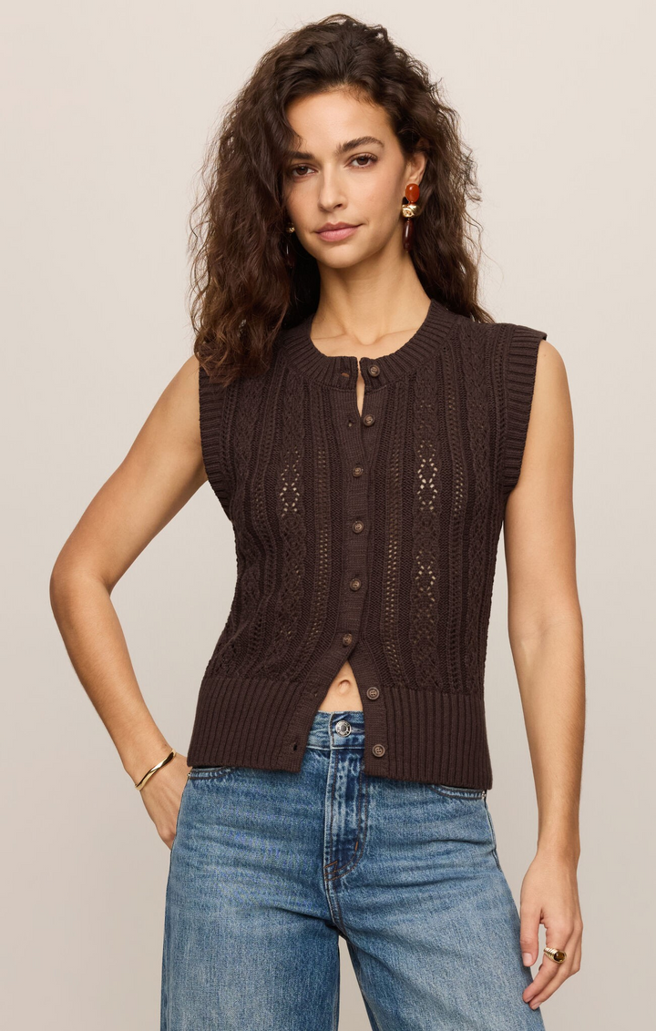 Woman wearing a brown knitted vest over a black top with blue jeans on a beige background