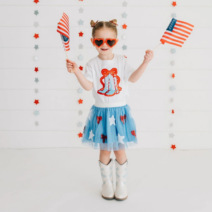 Child holding American flags against a decorative wall with stars and stripes wearing a patriotic tee shirt usa theme cowgirl boots