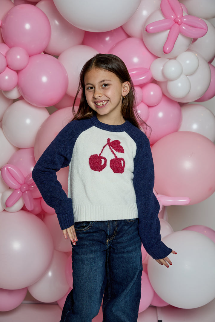 A young girl smiling in front of a pink and white balloon wall, wearing a navy blue and white raglan sweater with a red cherry graphic on the chest, paired with dark blue jeans.