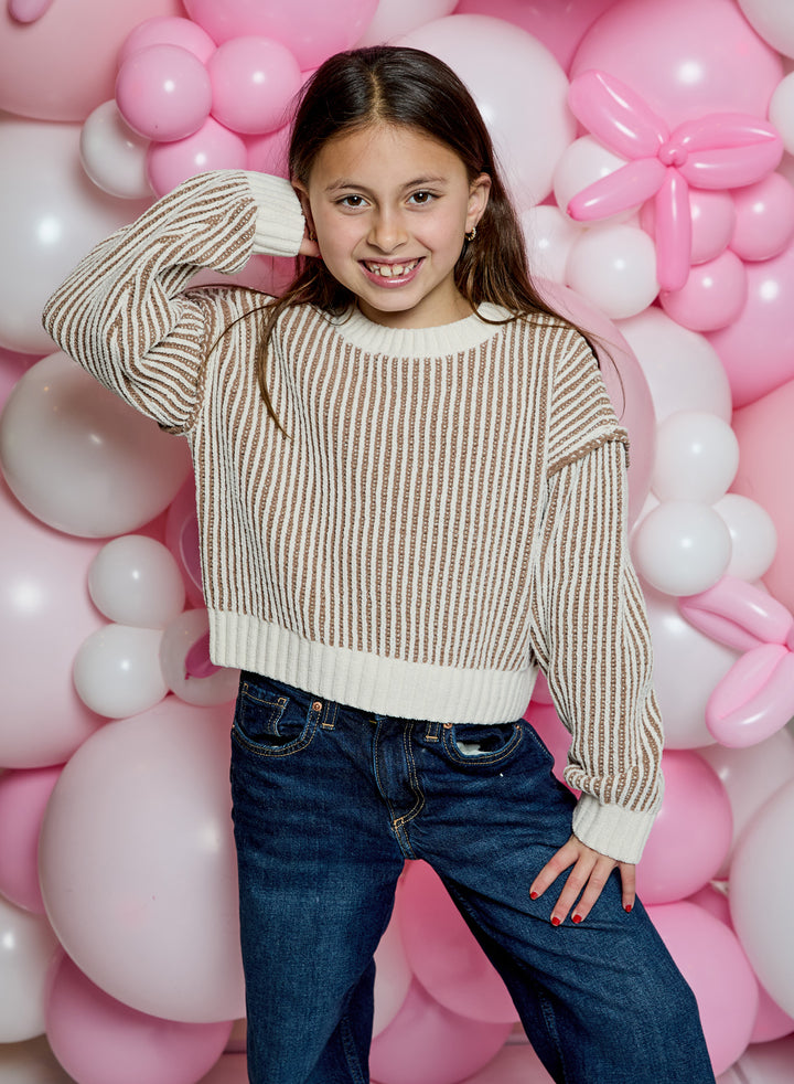 A young girl with long brown hair smiling in front of a pink and white balloon backdrop. She is wearing a beige and white vertically striped cropped knit sweater and dark blue jeans.