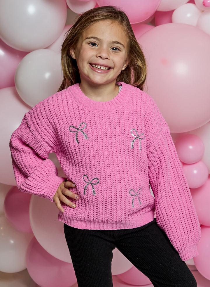 A young girl smiling and posing in a bright pink knit sweater with embroidered bow details, set against a background of pink and white balloons.