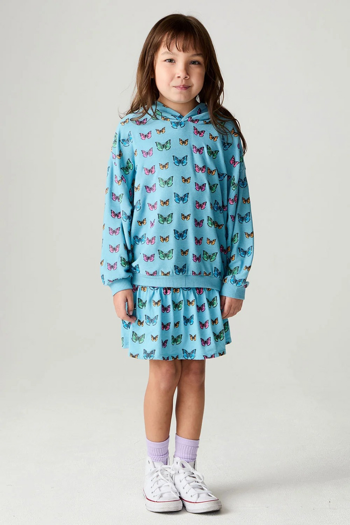 A young girl stands against a light gray studio background wearing a light blue hooded sweatshirt and matching pleated skirt set, both covered in a colorful butterfly print. She is also wearing light purple socks and white sneakers.