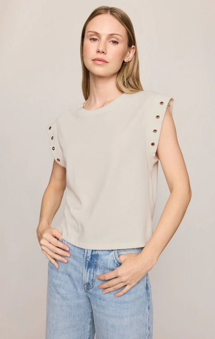 A woman modeling a light beige sleeveless top with decorative gold-tone eyelet details on the shoulders, paired with light-wash blue jeans, against a plain light gray studio background.