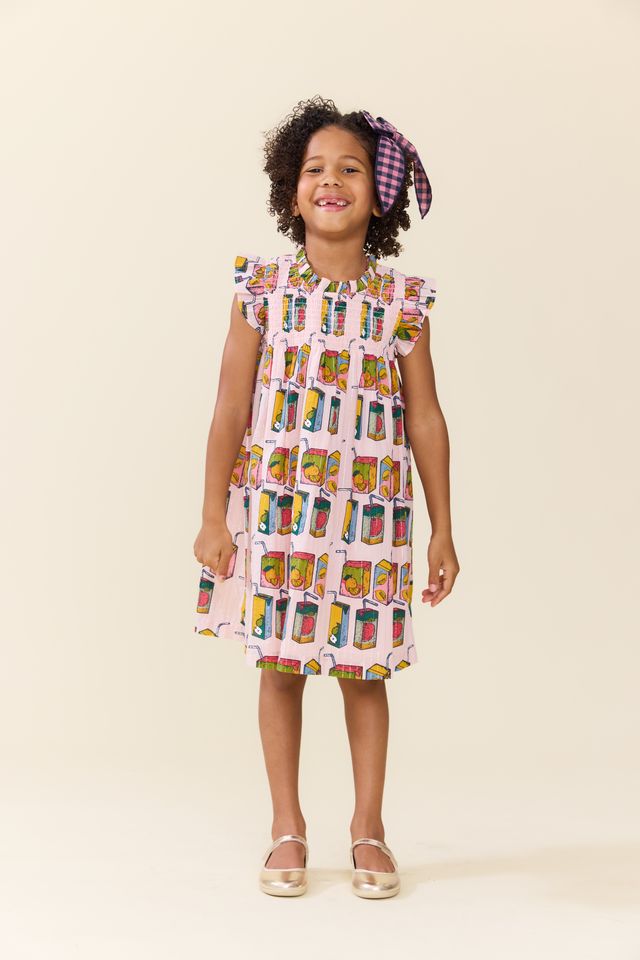 A young girl with curly hair wearing a pink dress with a colorful juice box pattern and a purple plaid headband, standing against a solid beige background. pink chicken