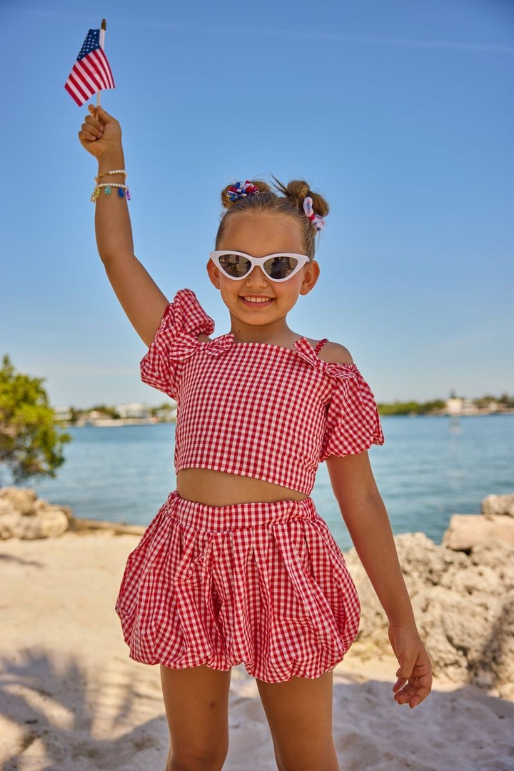 Child in a red checkered outfit holding an American flag on a beach. Lola & the boys 4th of july