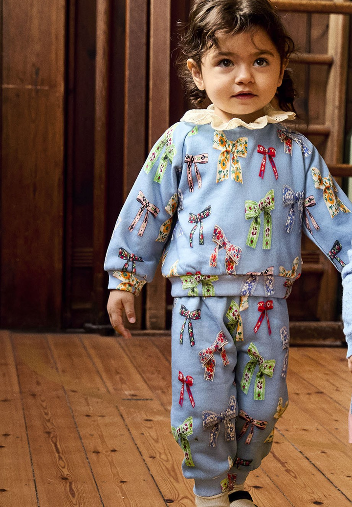 Two children in matching outfits standing on a wooden floor with wooden crates in the background. MOLO baby set bows